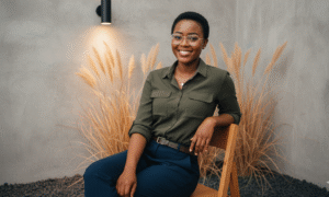 Create an image based on the attached original photo. The face, eyes, nose, mouth, and figure must be 100% identical to the attached original. Create a realistic portrait (face must remain 100% the same, do not modify). ​Hair: Same hairstyle, natural straight hair. ​Makeup: Natural tone makeup, faint rosy cheeks, eyes look big and bright, thin eyeliner, rosy pink lips. ​Outfit: Wearing the outfit from the second attached image. ​Glasses: Thin gold-rimmed square glasses. ​Scene: The background is a grey/bare concrete wall with a black cylindrical wall lamp shining downwards. Behind the subject, there are decorative plants resembling dry vetiver grass or reeds in a light brown or straw color. The floor has black and grey gravel/pebbles, looking like a photo corner in a coffee shop or café. ​Pose: A semi-reclined sitting pose, using the left hand to prop up the edge of the seat, the right leg slightly crossed forward. The body is slightly tilted, and the face is turned slightly to the left, looking at the camera with a beautiful, happy smile. ​Image Tone: Bright, warm, and comfortable tone (Earthy Tone), emphasizing white, beige, and light brown colors. The soft orange light provides a relaxing feeling and adds depth to the image. ​It should be a stylish and modern photograph, reflecting a tasteful relaxation in a beautifully decorated café atmosphere